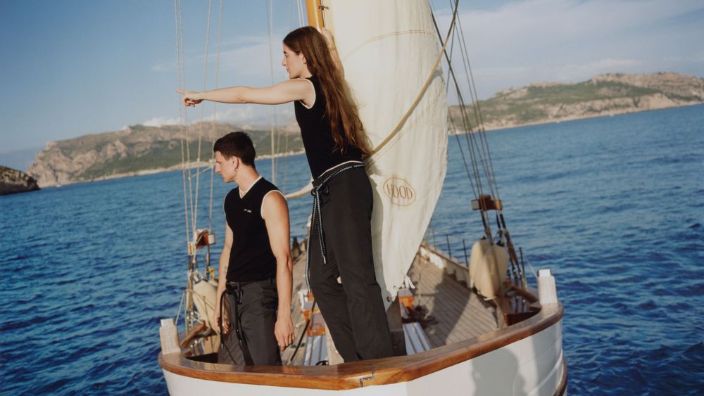 Male and female model in black organic cotton ensembles aboard a sailboat for the Yoox x Coco Capitán deadstock capsule campaign.