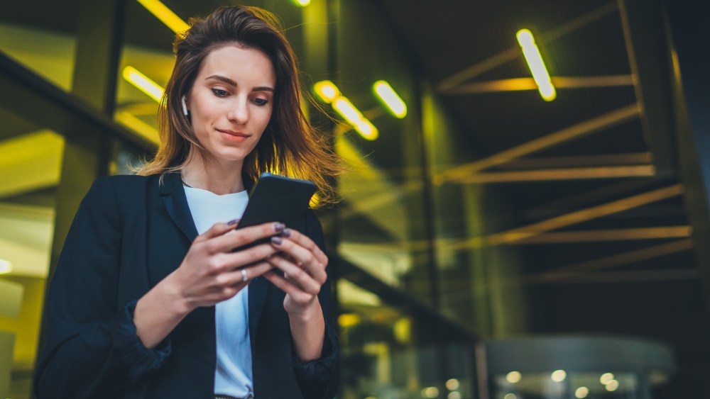 Successful female banker using smartphone outdoors while standing near his office background yellow neon lights, young woman professional manager working on mobile device near skyscraper at night