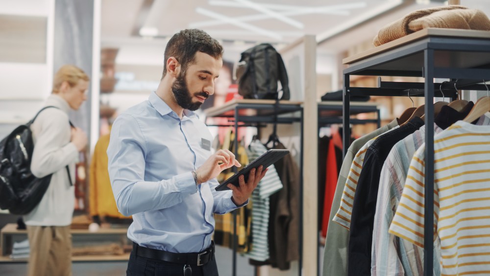 Clothing Store: Male Visual Merchandising Professional Uses Tablet Computer To Create Collection. Fashionable Shop Sales Retail Manager Checks Stock. Small Business Owner Orders Stylish Items