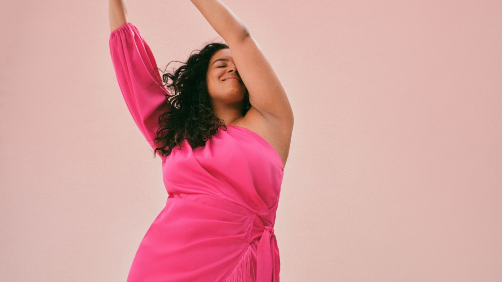 A fuchsia one-shoulder dress from Eloquii shown on a survey model with her arms in a striking overhead pose, as the brand makes its way to rental platform Nuuly.