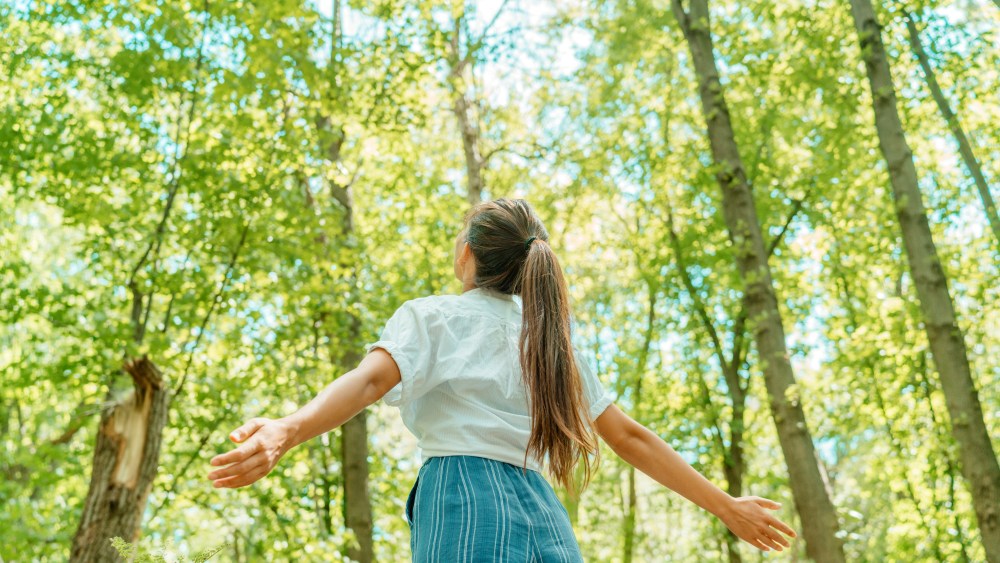 Free woman breathing clean air in nature forest. Happy girl from the back with open arms in happiness. Fresh outdoor woods, wellness healthy lifestyle concept.