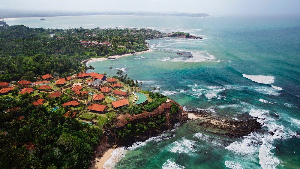 Cape Weligama, Sri Lanka features an aerial view of turquoise seas hitting up against a lush green coast peppered with red-roofed houses.