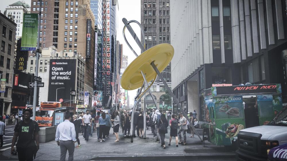 The Garment District's Big Button – a 28-feet tall yellow statue of button and needle – is stationed on Seventh Ave at 39th Street. In background, people walk by in crowds.