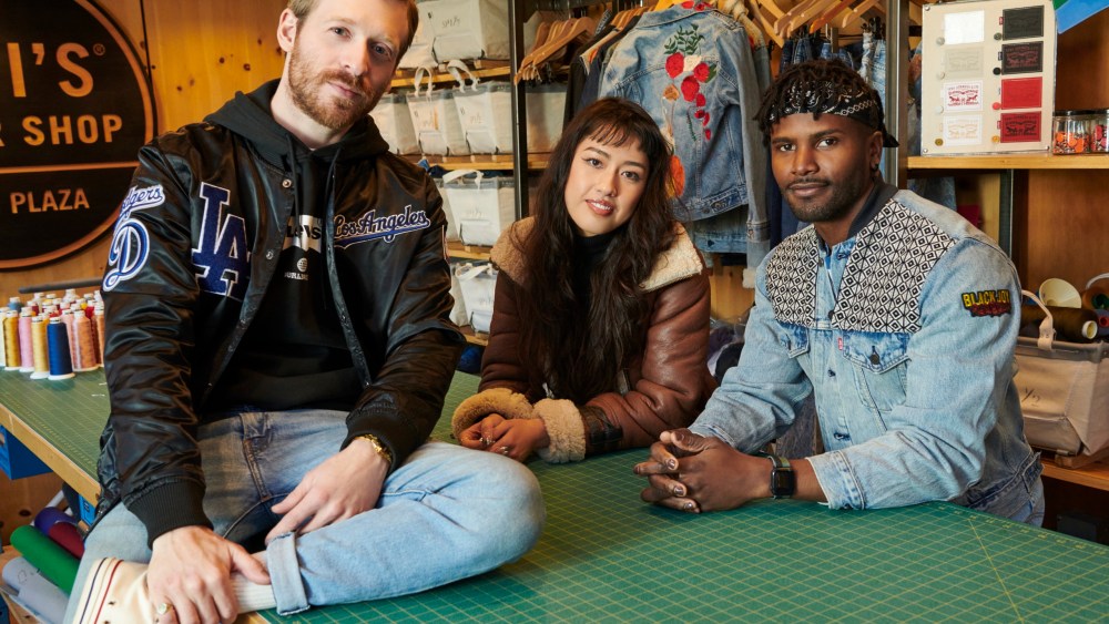 Three people of different ethnicities sit in a fashion work room wearing denim, looking at the camera.