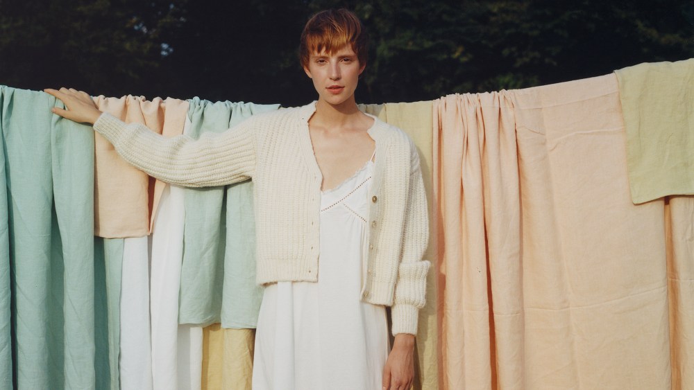 A woman wearing a white dress and cream cardigan stands in front of colorful sheets hanging on a line outdoors.