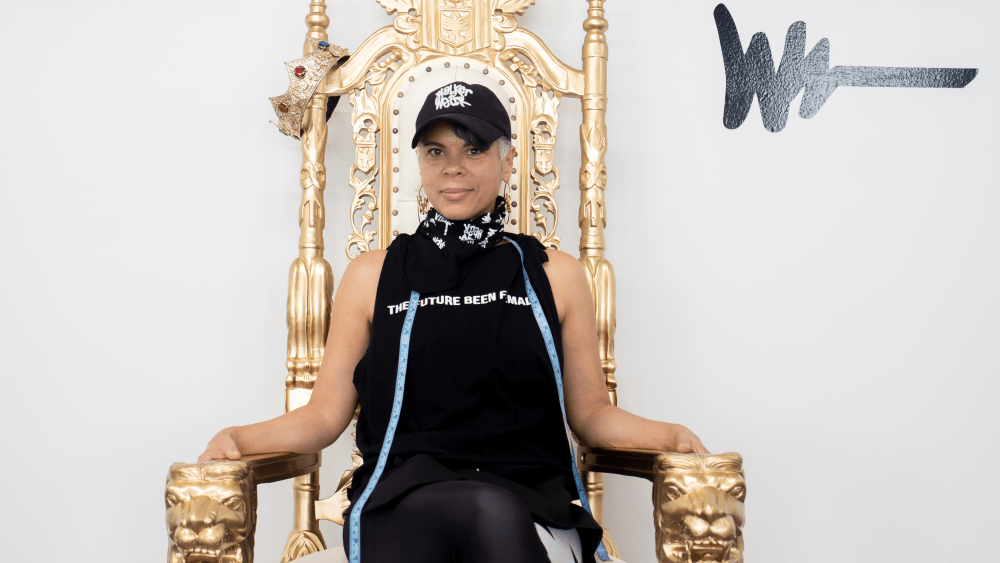 April Walker sits on a large throne-like chair under a sign that reads "The Future Been Female" in black letters.