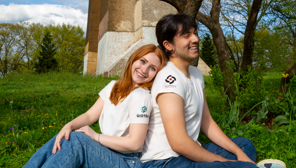Two people sitting back to back under a tree in the grass, smiling and wearing white recycled T-shirts with blue jeans.