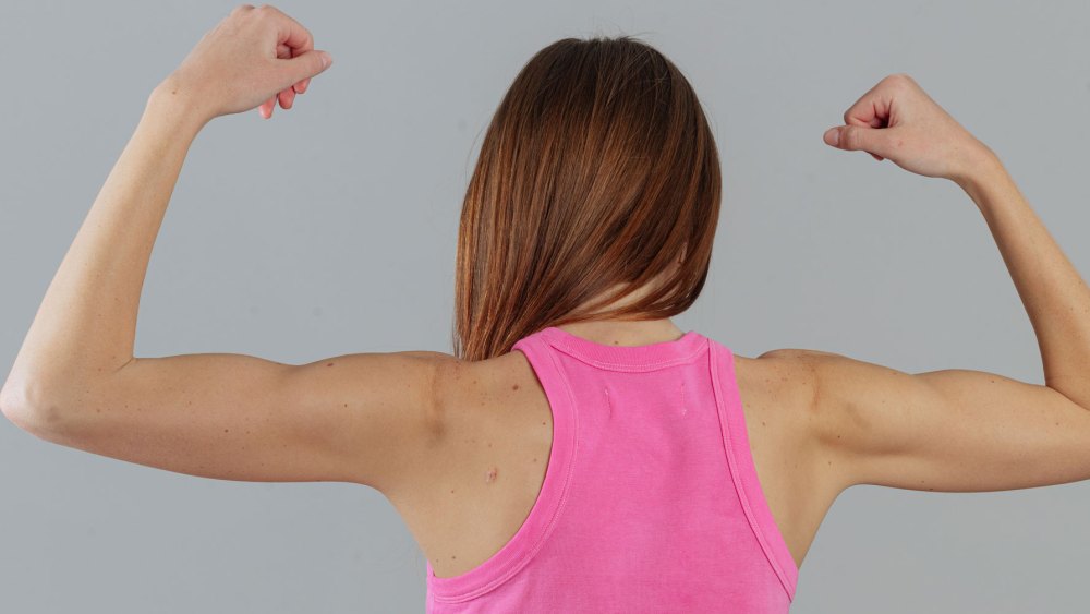 From back, a tanned model flexes her biceps wearing hot pink fascia tank and underwear from The Big Favorite.