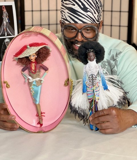 Designer Byron Lars poses with Sugar from his Chapeaux collection for Barbie on the left. She sits in a pink hat box with a hat covering her reddish big curly hair. She is wearing a blue satin skirt and red pumps. On the right is Mbili from his Treasures of Africa collection. She has a big curly afro and is wearing a blue and white outfit that includes feathers.