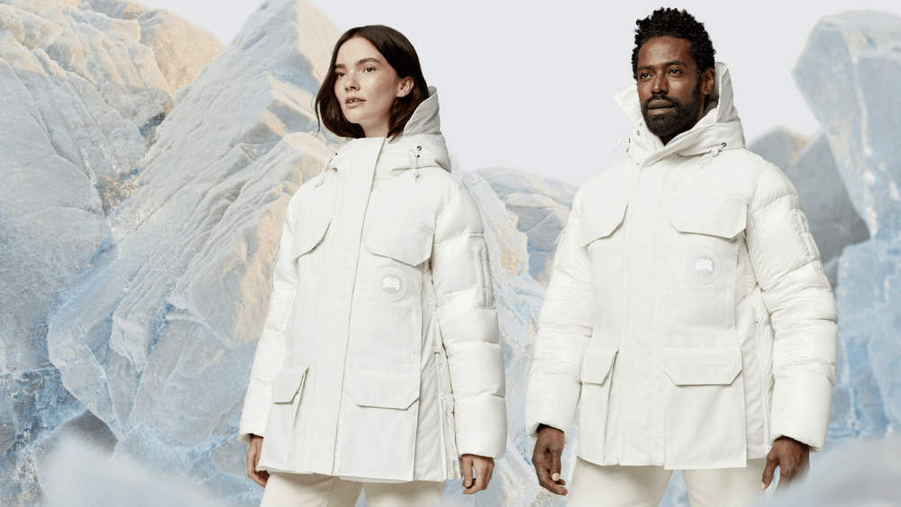 A snowy mountainous backdrop with two models posed in white Canada Goose jackets.