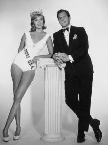 June 1965:  Bobbi Johnson, Miss USA 1964, and American actor and singer Pat Boone lean against a pillar in a full-length promotional portrait for the 'Miss USA 1965 Beauty Pageant,' in which Boone and Johnson appeared. Johnson is wearing a crown and a sash over a bathing suit.  (Photo by CBS Photo Archive/Getty Images)