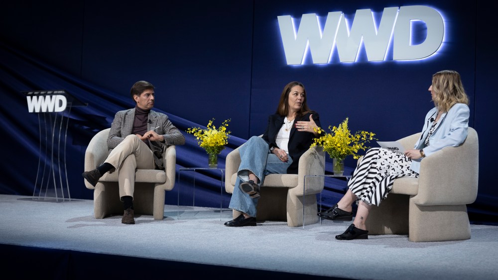 On stage in front of a blue backdrop with large white letters reading WWD: Stefano Canali, President & Chief Executive Officer, Canali Group and Amy Williams, Chief Executive Officer, Citizens of Humanity Group with Kaley Roshitsh, Editorial Director, Sustainable Apparel Coalition at the 2023 WWD Apparel & Retail CEO Summit.