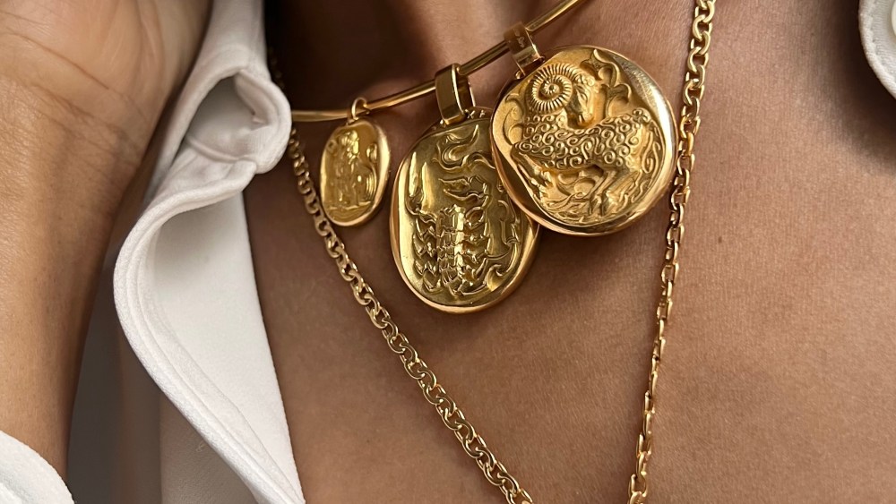 Close up of a woman, her neck adorned with two different gold chains, one featuring three zodiac pendants, the other featuring two zodiac pendants.