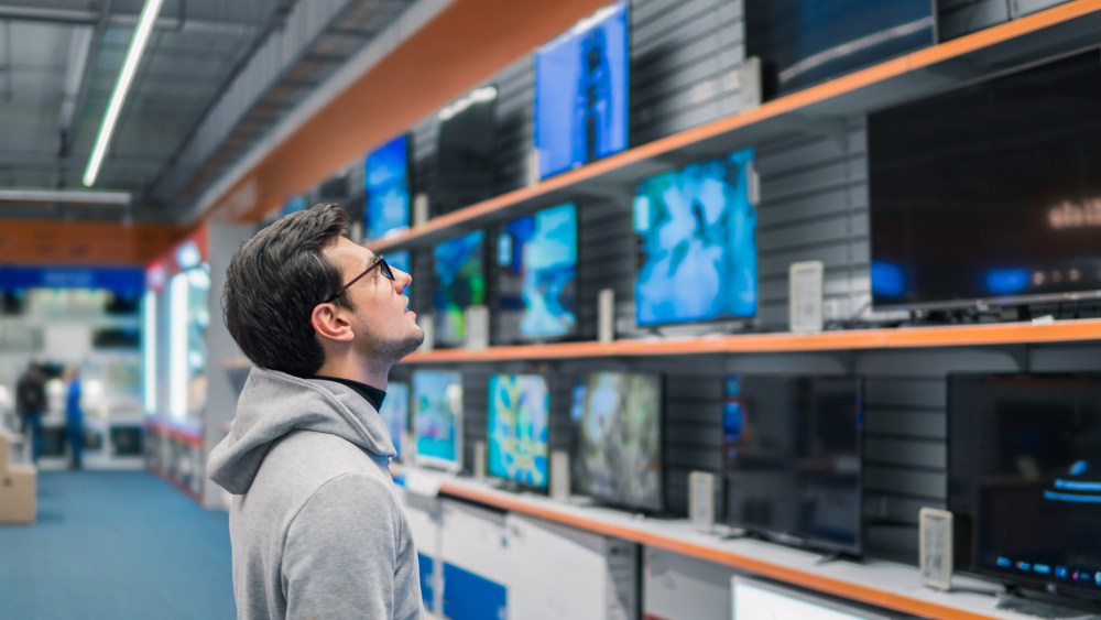 Smart modern male customer choosing large TV-sets at electronics store. He looks wondered. New screen generations.
