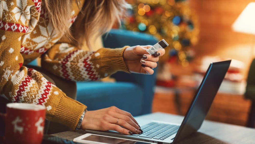 Woman doing online shopping at Christmas: she is sitting on the sofa at home, connecting with her laptop and holding a credit card, Christmas tree in the background