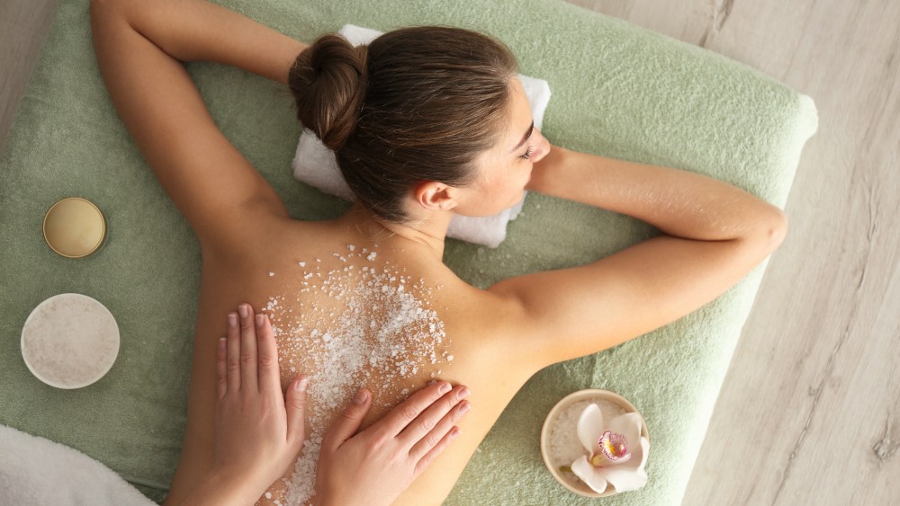 Young woman having body scrubbing procedure with sea salt in spa salon, top view