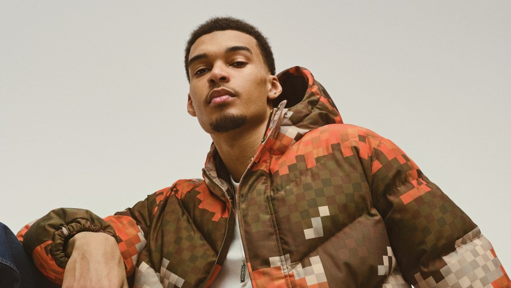 Victor Wembanyama in a Louis Vuitton puffer jacket with a pixelated pattern sitting on a Louis Vuitton monogram trunk