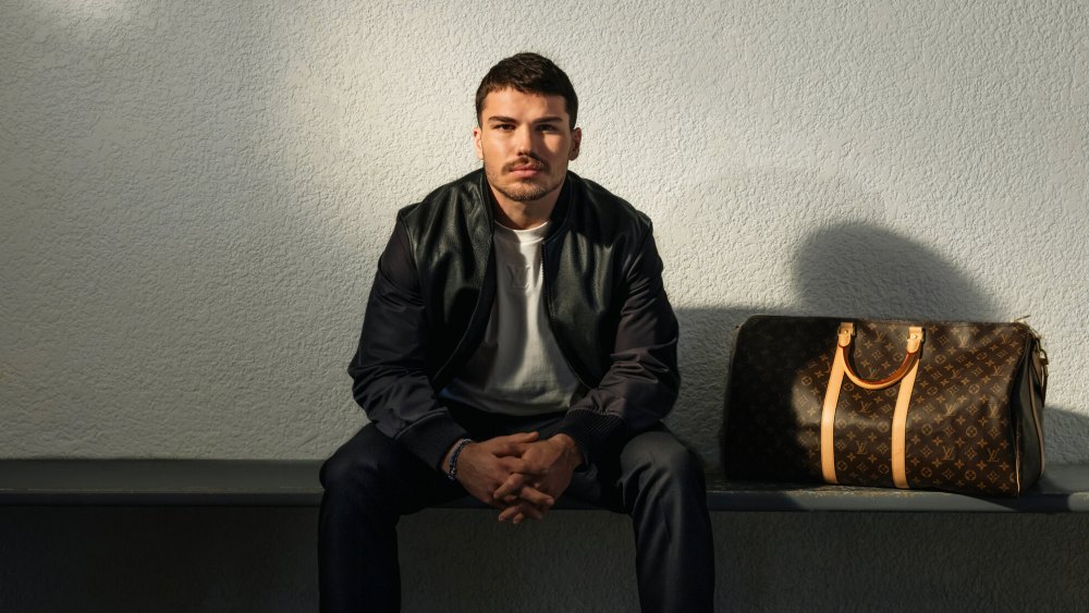 Rugby player Antoine Dupont poses in a locker room next to a Louis Vuitton monogram Keepall bag.