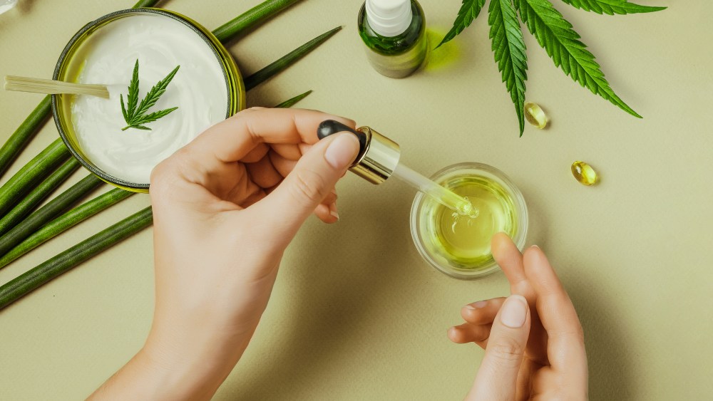 Pipette with CBD cosmetic oil in female hands on a table background with cosmetics, cream with cannabis and hemp leaves, marijuana. Flat lay, top view.