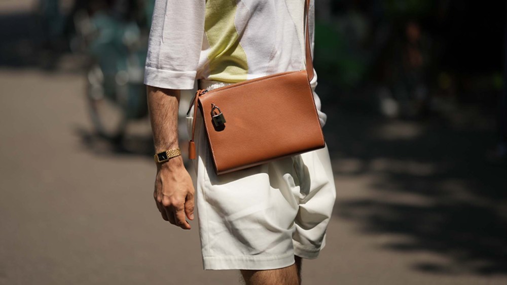 A man wears a pale gray with large pale khaki strap pattern t-shirt, a camel shiny leather crossbody bag, beige suit shorts, a gold watch