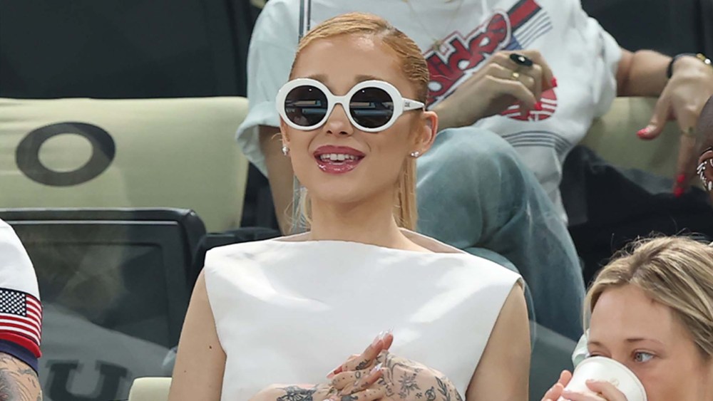 Ariana Grande sitting at a sporting event wearing a white dress and round Loewe white sunglasses