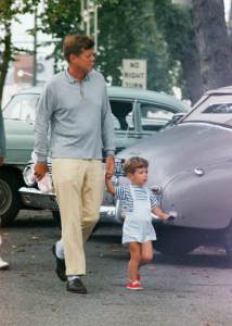 John F. Kennedy walks holding John Jr.'s hand at Hyannisport, Massachusetts. The president carries a pink teddy bear, and John Jr. carries a toy helicopter.