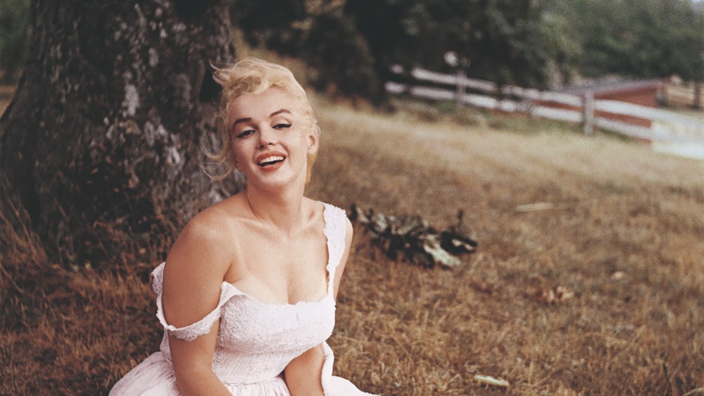 Marilyn Monroe sitting in the grass barefoot, wearing a pink dress in 1957 in Amagansett, New York.