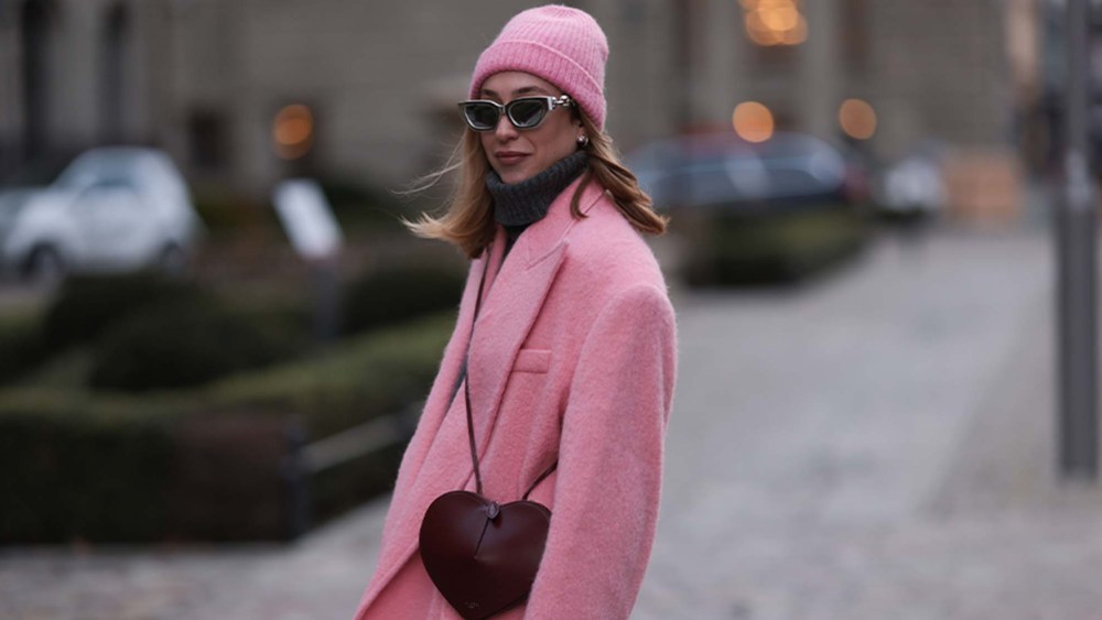woman wears a pink beanie hat, pink blazer, and heart-shaped handbag