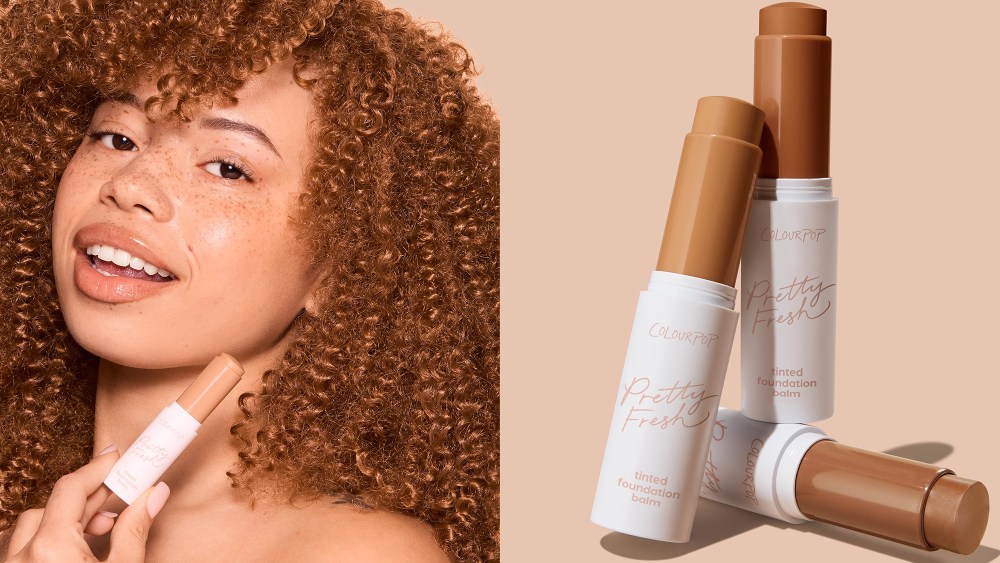 woman with coily and poofy hair holding up a foundation stick while smiling; three foundation sticks in various shades arranged artfully over a nude background