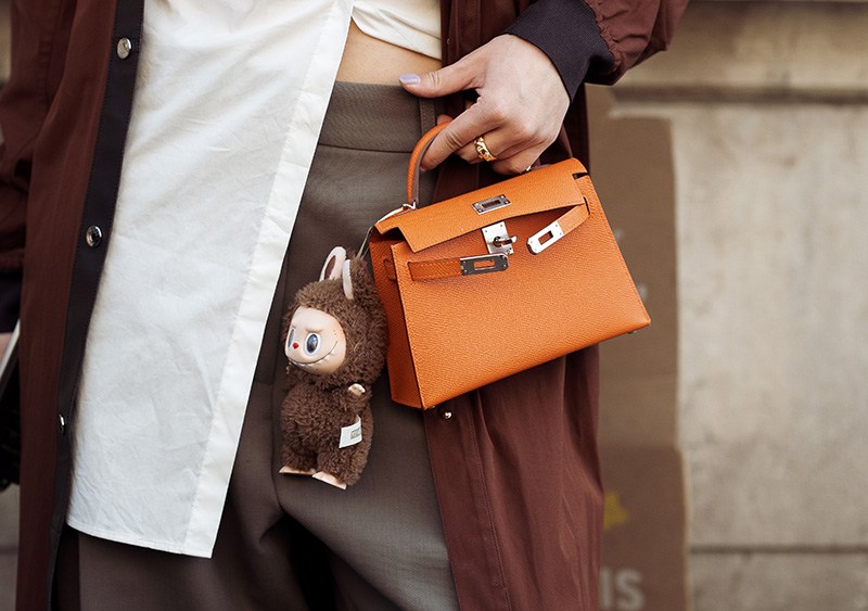 PARIS, FRANCE - MARCH 08: A guest wears brown coat, white blouse, orange Hermes bag with a bag charm brown Labubu monster, grey pants outside the Hermes fashion show during the Womenswear Fall/Winter 2025/2026 as part of Paris Fashion Week on March 08, 2025 in Paris, France. (Photo by Raimonda Kulikauskiene/Getty Images)