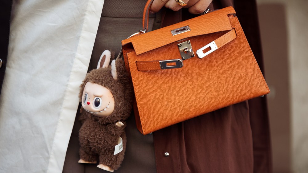PARIS, FRANCE - MARCH 08: A guest wears brown coat, white blouse, orange Hermes bag with a bag charm brown Labubu monster, grey pants outside the Hermes fashion show during the Womenswear Fall/Winter 2025/2026 as part of Paris Fashion Week on March 08, 2025 in Paris, France. (Photo by Raimonda Kulikauskiene/Getty Images)