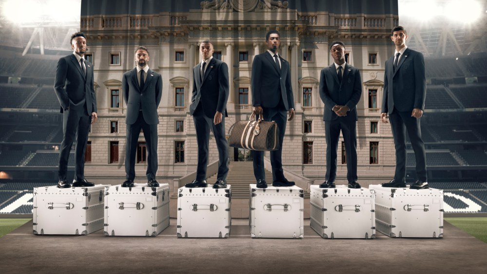 Éder Militão, Daniel Carvajal, Kylian Mbappé, Jude Bellingham, Vinicius Júnior and Thibaut Courtois standing on Louis Vuitton trunks, wearing the LV x Real Madrid formal wear collection.