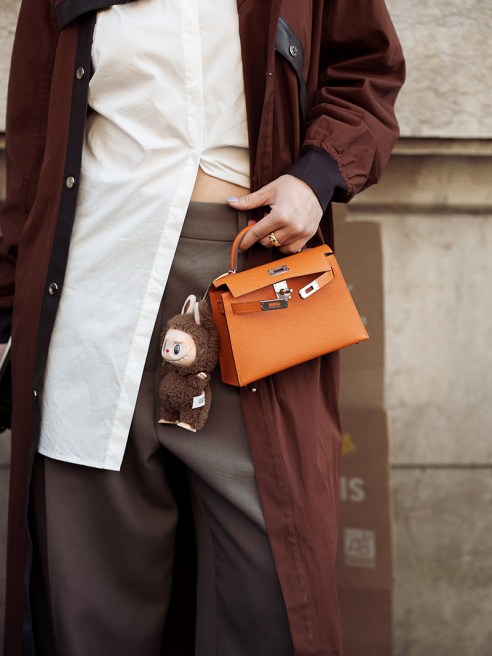 A guest wears brown coat, white blouse, orange Hermes bag with a bag charm brown Labubu monster, grey pants outside the Hermes fashion show during the Womenswear Fall/Winter 2025/2026 as part of Paris Fashion Week on March 08, 2025 in Paris, France.