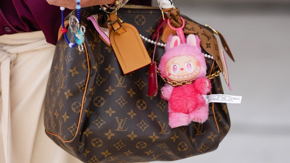 PARIS, FRANCE - JULY 08: A guest wears a loose-fitting, deep burgundy button-up shirt with the sleeves casually rolled up. The shirt is paired with cream-colored, wide-legged trousers featuring a tied waist detail. Accessories include a brown monogrammed Speedy handbag from Louis Vuitton adorned with colorful keychains and a fluffy pink Labubu charms. Footwear consists of white sneakers from Gucci with visible wear, outside Chanel, during Paris Fashion Week - Haute Couture Fall/Winter 2025/2026, on July 08, 2025 in Paris, France (Photo by Edward Berthelot/Getty Images)