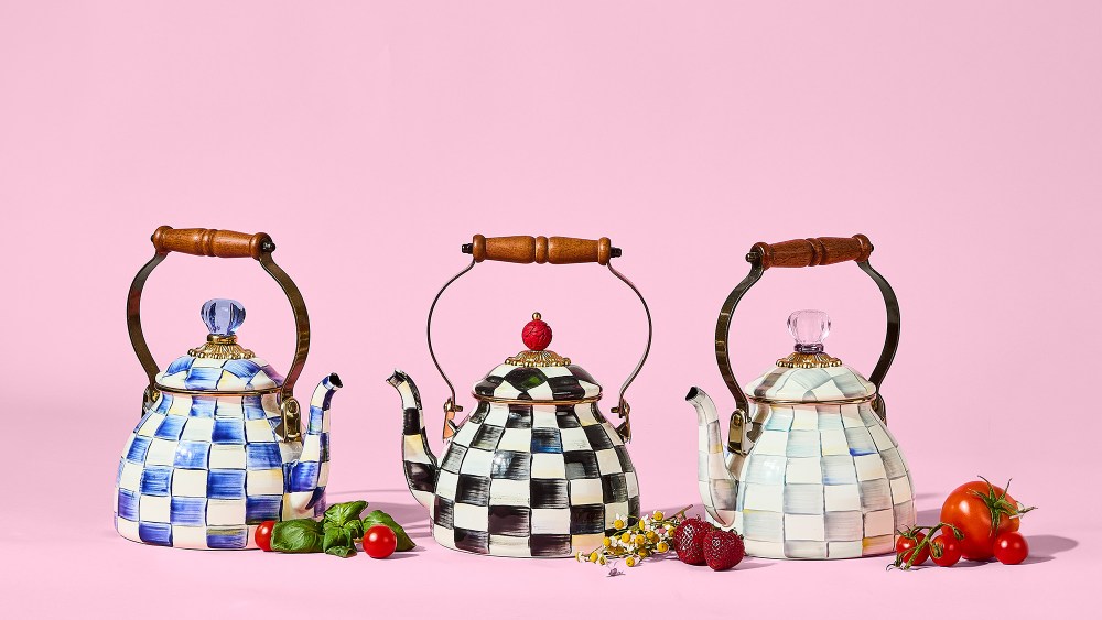 three checkered tea kettles with wood-topped handles resting next to an assortment of fruits and vegetables, in front of a pink backdrop