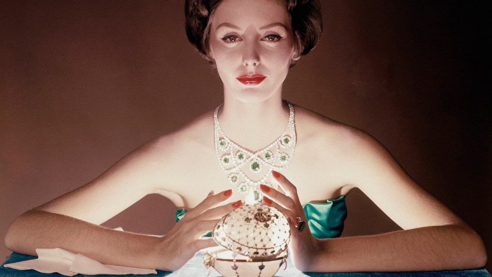 Seated model illuminated by light from Faberge egg, wearing lacy collar of square-cut emeralds and round diamonds.