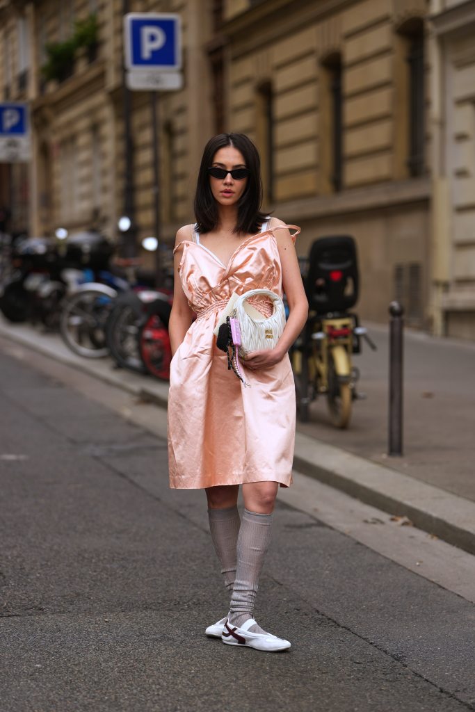 PARIS, FRANCE - OCTOBER 06: A guest wears shoulder-length straight dark brown hair with a center part, black narrow cat-eye sunglasses, a white ruched leather shoulder bag from Miu Miu with attached keychain charms including a pink fob and a black pom-pom, a white underlayer tank strap visible under a peach metallic satin knee-length dress with thin off-the-shoulder straps, a V-neckline, gathered elastic waist and side pockets, knee-high ribbed taupe socks, white Mary Jane shoes with flat soles, outside Miu Miu, during Paris Fashion Week - Womenswear Spring Summer 2026, on October 06, 2025 in Paris, France (Photo by Edward Berthelot/Getty Images)
