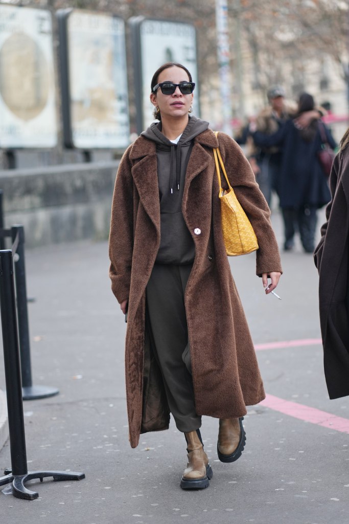 PARIS, FRANCE - JANUARY 20: A guest wears dark brown hair pulled back with a center part, black rectangular sunglasses, metal hoop earrings, a yellow coated canvas shoulder bag with an allover embossed monogram pattern from Goyard, a brown teddy faux fur long coat with notch lapels, a dark gray hooded sweatshirt and matching dark gray jogger pants, brown leather Chelsea boots shoes with black lug soles, outside 424, during Paris Fashion Week - Menswear Fall/Winter 2026-2027, on January 20, 2026 in Paris, France (Photo by Edward Berthelot/Getty Images)