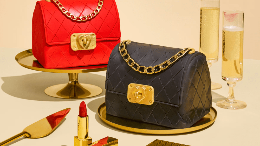 two handbag-shaped cakes in red and black standing on cake trays next to lipstick, a serving spoon, and two full champagne flutes