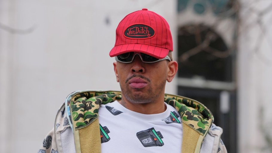 A guest wears a red baseball cap with a black oval logo patch from Von Dutch, a beige hooded zip jacket with an allover black graphic print and a green camouflage lining, a white printed T-shirt, outside Rick Owens, during Paris Fashion Week - Menswear Fall/Winter 2026-2027, on January 22, 2026 in Paris, France.