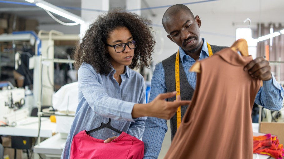 Two diverse adult fashion designers, a man and a woman, are discussing clothing designs and fabrics, holding garments in a textile production workshop, highlighting teamwork in the garment industry